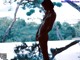 A naked woman standing in front of a pool with trees in the background.